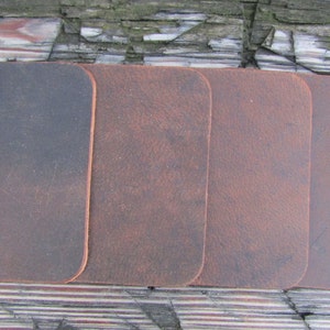 May include: Four leather coasters in different shades of brown. The coasters are rectangular and have a rustic, distressed look.