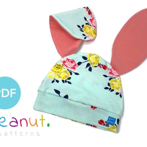 May include: A light blue hat with a floral pattern and two coral pink bunny ears. The hat has a small blue label with the text "from peanut patterns".