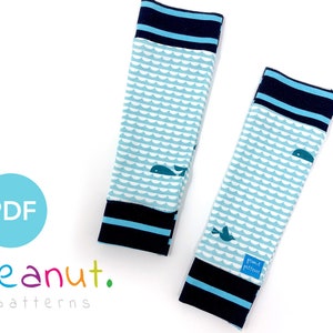 May include: A pair of blue and white leg warmers with a whale and wave pattern. The leg warmers have a navy blue cuff with a white stripe. The leg warmers are made by Peanut Patterns.