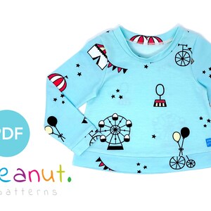May include: A light blue long-sleeved shirt with a circus theme print. The print features a Ferris wheel, balloons, a bicycle, a tent, and stars. The shirt has a round neckline and long sleeves.