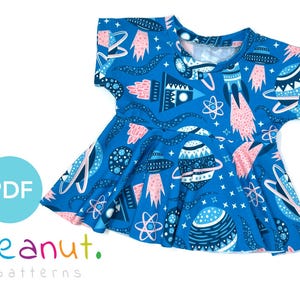 Puede incluir: Un vestido para niños de color azul y rosa con un estampado de temática espacial. El vestido tiene mangas cortas y una falda fruncida. El estampado presenta planetas, cohetes, estrellas y átomos.