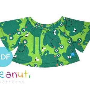 May include: A green t-shirt with a frog print. The shirt has a round neckline and short, wide sleeves. The fabric has a green background with white and black frogs and flies.