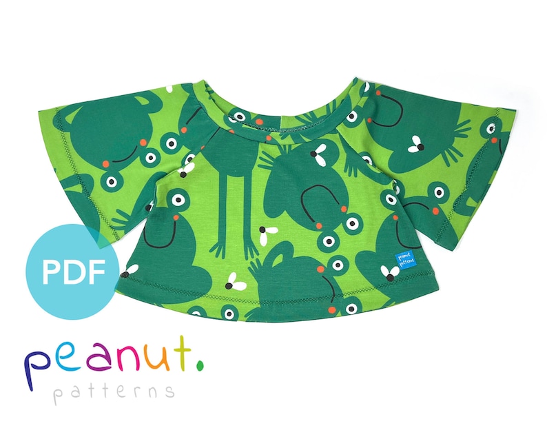 May include: A green t-shirt with a frog print. The shirt has a round neckline and short, wide sleeves. The fabric has a green background with white and black frogs and flies.