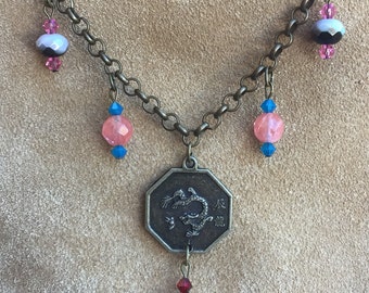 May include: Antique bronze-toned necklace featuring a dragon pendant and various beaded accents. The necklace has a chain with pink, blue, and iridescent beads. The pendant is an octagon shape with a dragon design and Chinese characters.