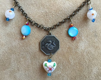 May include: A bronze-toned necklace featuring a chain with decorative elements. The necklace includes a central octagonal pendant with a snake design and Chinese characters. It also has blue and white beads, and a heart-shaped pendant with floral details.