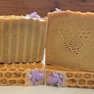 May include: Four bars of honey soap with a honeycomb design and purple bee decorations. The soap is a light brown color.