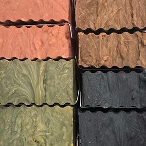May include: A close-up shot of several bars of handmade soap in various colors. The soaps are arranged in rows and include shades of pink, brown, green, and black. Each bar has a wavy top edge and a textured appearance.
