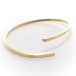 May include: A gold-toned metal bangle bracelet with a squared-off design. The bracelet is open and has a simple, minimalist style.