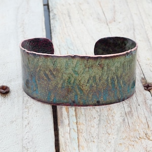 May include: A wide cuff bracelet featuring a textured surface with a patina finish. The metal displays a gradient of colors including deep green, oxidized blue, and hints of dark copper along the edges and the interior rim.