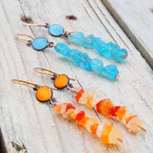 May include: Two pairs of handmade earrings. One pair features blue enamel circles with blue crystal beads. The other pair has orange enamel circles with orange and white crystal beads. Both pairs have gold-colored ear wires.