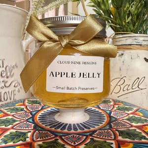 Può includere: Un piccolo barattolo di gelatina di mele con un fiocco dorato. L'etichetta recita "APPLE JELLY -Small Batch Preserves-" e "CLOUD NINE DESIGNS". Il barattolo è su un piedistallo bianco su un piatto fantasia. Altri barattoli e fiori sono sullo sfondo.