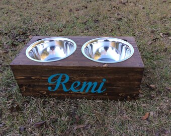 May include: A wooden dog bowl stand with two stainless steel bowls. The stand is stained dark brown and has the name "Remi" written in teal blue lettering.
