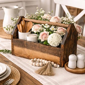 May include: A wooden toolbox filled with pink roses, white daisies, and greenery. The toolbox also contains wooden spoons, candles, and a white ceramic container. A wooden bead garland and salt and pepper shakers are on the table.