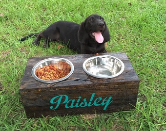 May include: A wooden dog bowl stand with two stainless steel bowls. The stand is painted dark brown and has the name "Paisley" written in teal paint. A black dog is sitting in front of the stand.