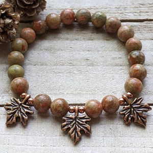 May include: A beaded bracelet featuring round, earthy-toned beads in shades of brown, green, and red. The bracelet is adorned with three copper-colored maple leaf charms. The bracelet is displayed on a light-colored wooden surface.