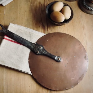 May include: A round copper lid with a dark, weathered iron handle featuring a circular loop at the end. The handle is attached to the copper disc with three visible rivets. The lid rests partially on a folded white cloth with a red stripe.