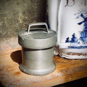 May include: A small, antique pewter container with a handle on top, set on a wooden surface. The container has a cylindrical shape with a lid and etched horizontal lines. A decorative white and blue pitcher is in the background.