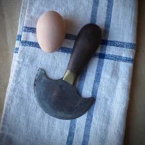 May include: A rustic kitchen scene featuring a brown egg and a mezzaluna knife with a wooden handle and brass ferrule, resting on a white and blue striped linen towel. The knife blade has a curved shape.