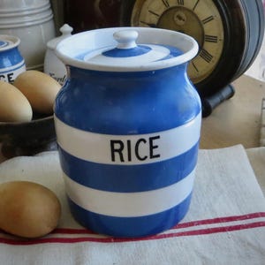 May include: A cylindrical ceramic storage jar with a lid, featuring horizontal blue and white stripes. The word RICE is printed in black capital letters on the front of the jar. The jar has a smooth, glossy finish and a rounded rim.
