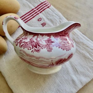 May include: Creamer with a white base and a red floral pattern. The creamer has a handle and a spout. The creamer is sitting on a white cloth with red stripes. Three brown eggs are in the background.