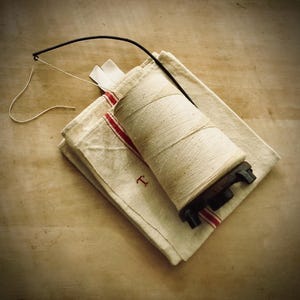 May include: A spool of natural twine sits atop a folded linen cloth with red stripes and the letter "T". A black wire thread guide extends from the spool. The items are arranged on a light wooden surface.