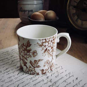 May include: A white ceramic mug with a brown floral pattern, featuring a handle and a wide opening. The mug is placed on a vintage paper with handwritten text. In the background, there's a jar, eggs, and a clock.