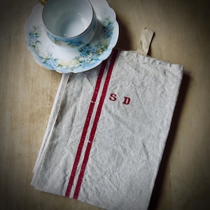 May include: A vintage teacup and saucer set with a floral design, alongside a folded linen towel. The towel is off-white with two red stripes and the initials "SD" embroidered in red. The teacup is empty.