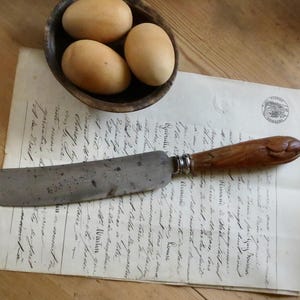 May include: An antique knife with a wooden handle rests on a vintage letter. Three brown eggs sit in a small wooden bowl. The scene is set on a wooden surface, evoking a rustic, historical feel.