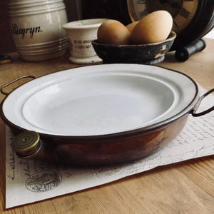 May include: A round, shallow copper pan with a white enamel interior and two thin metal side handles. The pan features a small, circular brass-colored cap attached to the side rim, providing a distinct contrast to the metallic copper exterior.