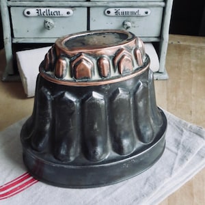 May include: Antique metal fluted mold with a copper rim. The mold is dark gray with a decorative design and sits on a gray and white striped cloth. The background includes a vintage cabinet with labeled drawers.