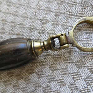 May include: Close-up of a vintage bottle opener. The opener features a dark brown handle with brass accents and a brass ring. The handle is oval-shaped and appears to be made of wood or a similar material. The brass components show signs of age.