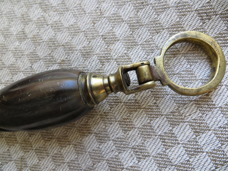 May include: Close-up of a vintage bottle opener. The opener features a dark brown handle with brass accents and a brass ring. The handle is oval-shaped and appears to be made of wood or a similar material. The brass components show signs of age.