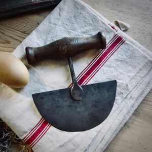 May include: Antique mezzaluna knife with a wooden handle and a curved blade. The knife rests on a white linen towel with red stripes. An egg is visible on the left side of the image. The knife is a kitchen utensil.
