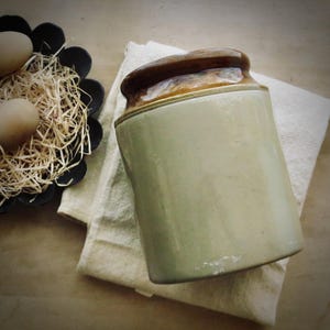 May include: A vintage, cylindrical stoneware jar with a brown lid and a light green body, resting on a cream-colored cloth. A black, flower-shaped bowl holds straw and two brown eggs, suggesting a rustic, farmhouse theme.