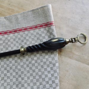 May include: A black and brass corkscrew with a twisted handle and a ring pull. The corkscrew rests on a gray and white checkered linen cloth with a red stripe.