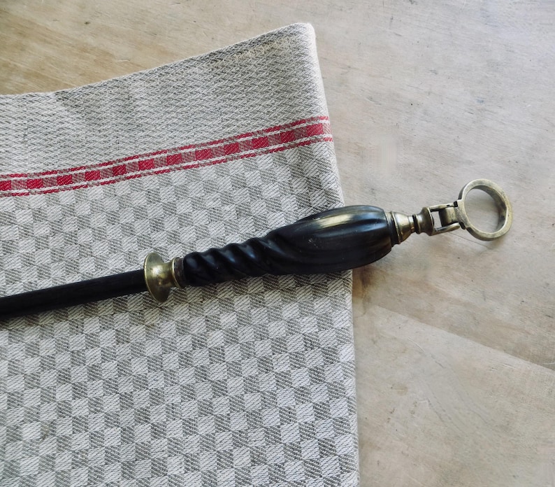 May include: A black and brass corkscrew with a twisted handle and a ring pull. The corkscrew rests on a gray and white checkered linen cloth with a red stripe.