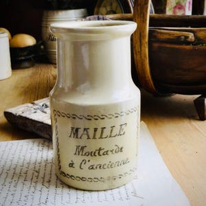 May include: A cream-colored ceramic mustard jar with a flared rim and a cylindrical body. The front features a decorative border enclosing the black printed text "MAILLE Moutarde à l'ancienne" in a serif font, centered on the vessel.