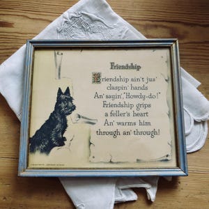 May include: Framed vintage print featuring a black Scottish Terrier dog illustration. The print has a blue frame and includes the poem "Friendship" with text in a decorative font. The print rests on a white, lacy cloth.