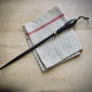 May include: A black sharpening steel with a decorative handle, resting on a folded gray and white checkered kitchen towel with red stripes. The handle has brass accents and a twisted design.