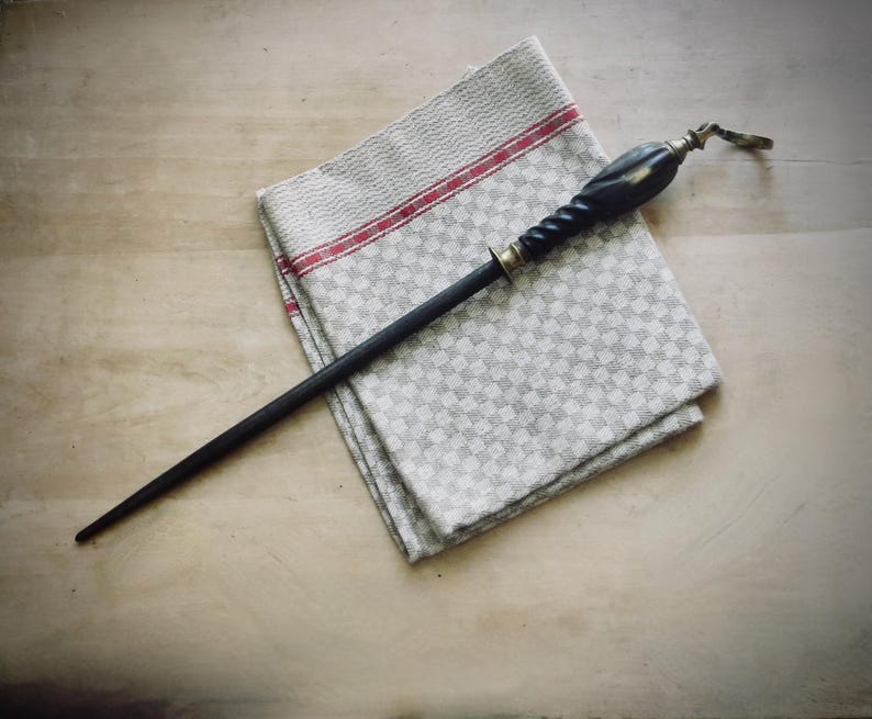 May include: A black sharpening steel with a decorative handle, resting on a folded gray and white checkered kitchen towel with red stripes. The handle has brass accents and a twisted design.