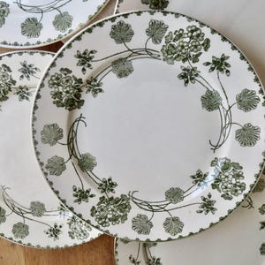 May include: A set of round ceramic plates featuring a cream-colored base with a dark green botanical pattern of flowers and leaves. The design includes a delicate floral border and a repeating vine motif that circles the center of each plate.