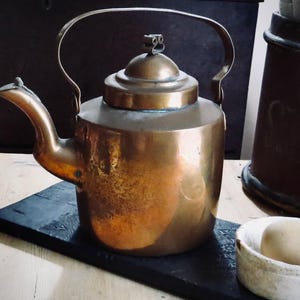 May include: A vintage copper kettle with a curved spout and a large handle. The kettle sits on a dark, rectangular trivet. The copper has a warm, aged patina, suggesting its use over time. A small, white bowl with a light-colored object is in the foreground.