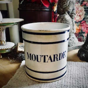 May include: A white ceramic container with the word "MOUTARDE" in blue lettering. The container has two blue stripes and a blue rim. The container is cylindrical and sits on a white woven surface.