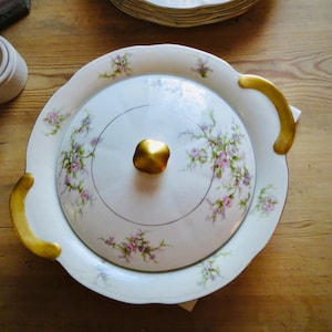 May include: A white porcelain serving dish with a lid, featuring a floral pattern in pink and green. The dish has gold-colored handles and a gold knob on the lid. A small white container is visible to the left.
