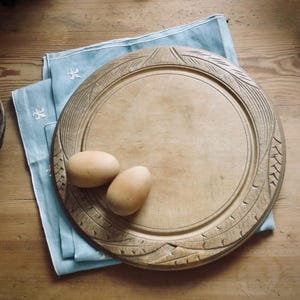 May include: A round wooden serving board with a carved border featuring linear and chevron patterns. Two smooth, light brown eggs rest on the surface of the board, which is placed atop a folded light blue cloth with white embroidered details.