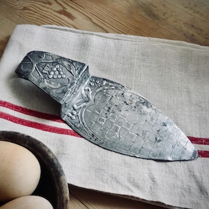 May include: An antique silver cake server with ornate floral and grape detailing. The server rests on a linen towel with red stripes, next to a wooden bowl with eggs. The server has a checkered pattern on the blade.