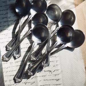May include: A collection of silver soup spoons with ornate handles, arranged on a vintage paper with handwritten script. The spoons have a reflective surface, catching the light and creating highlights. The overall aesthetic is classic and elegant.