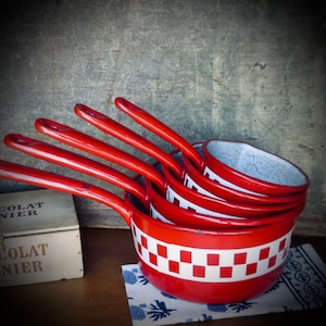 May include: A set of four red enamel saucepans with white checkered bands, stacked on top of each other. The saucepans are sitting on a white and blue patterned fabric. A wooden box with the text "Chocolat Menier" is visible in the background.
