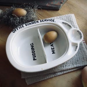 May include: A white ceramic Office Dinner Service plate with compartments for pudding and meat, plus sections for mustard, pickles, and salt. Two brown eggs are in the plate and in a nest. The plate rests on a gray cloth.