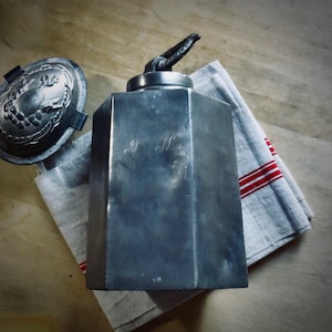 May include: A vintage metal container with a bird-shaped handle, next to a decorative lid. The container has engraved initials and sits on a gray and red striped cloth. The items are on a wooden surface.